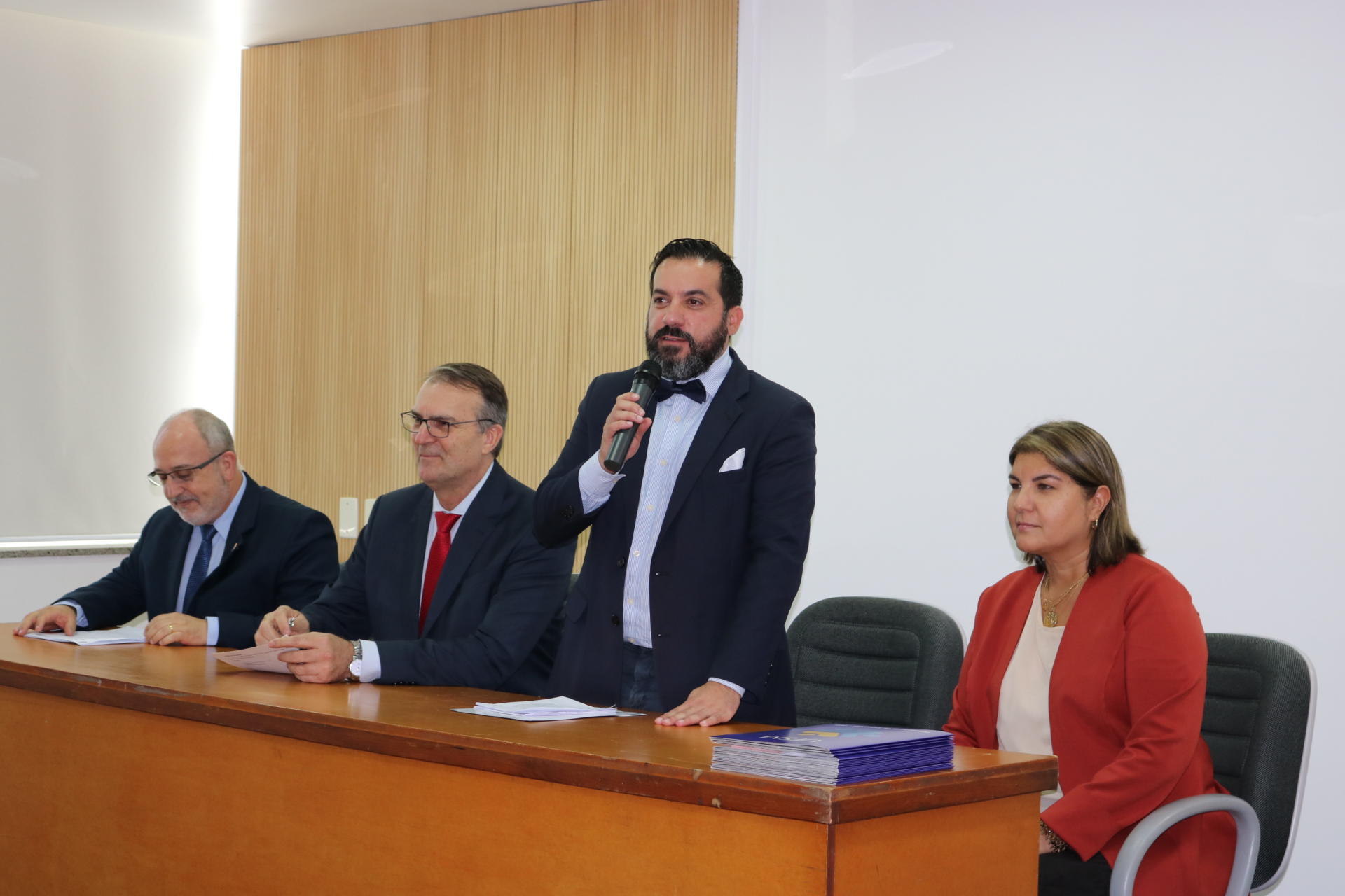 Três pessoas sentadas atrás da mesa e o pró-reitor de Extensão está em pé e segura o microfone.