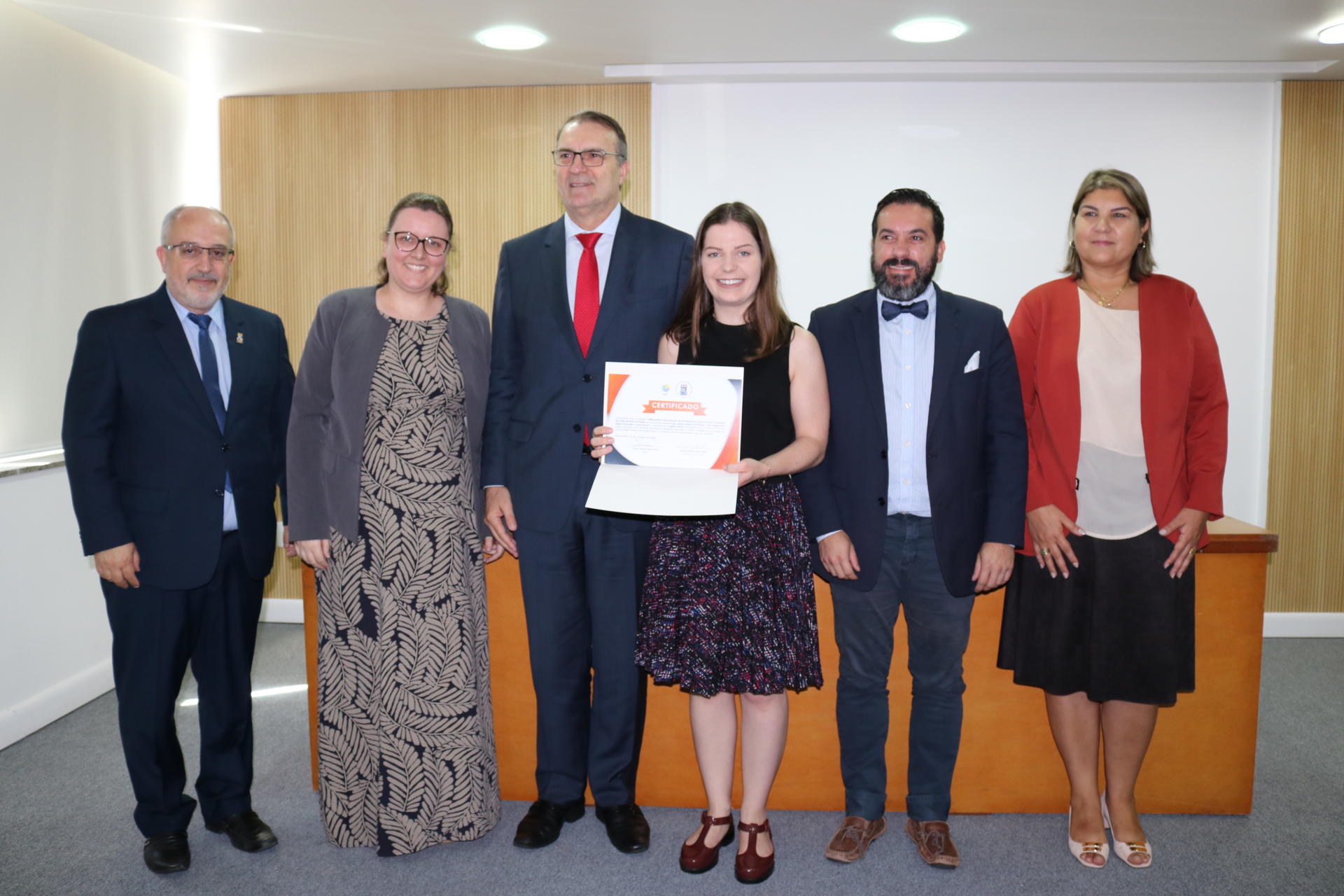 Seis pessoas em pé em frente do palco, incluindo o reitor e o pró-reitor de Extensão. Uma das alunas segura o certificado