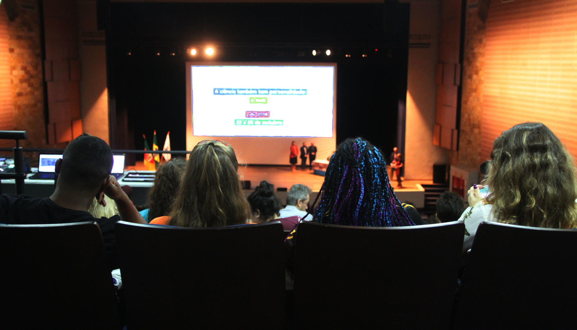Foto da perspectiva do público mostrando o Centro de Convenções lotado e com pessoas no palco e com telão com informações do evento