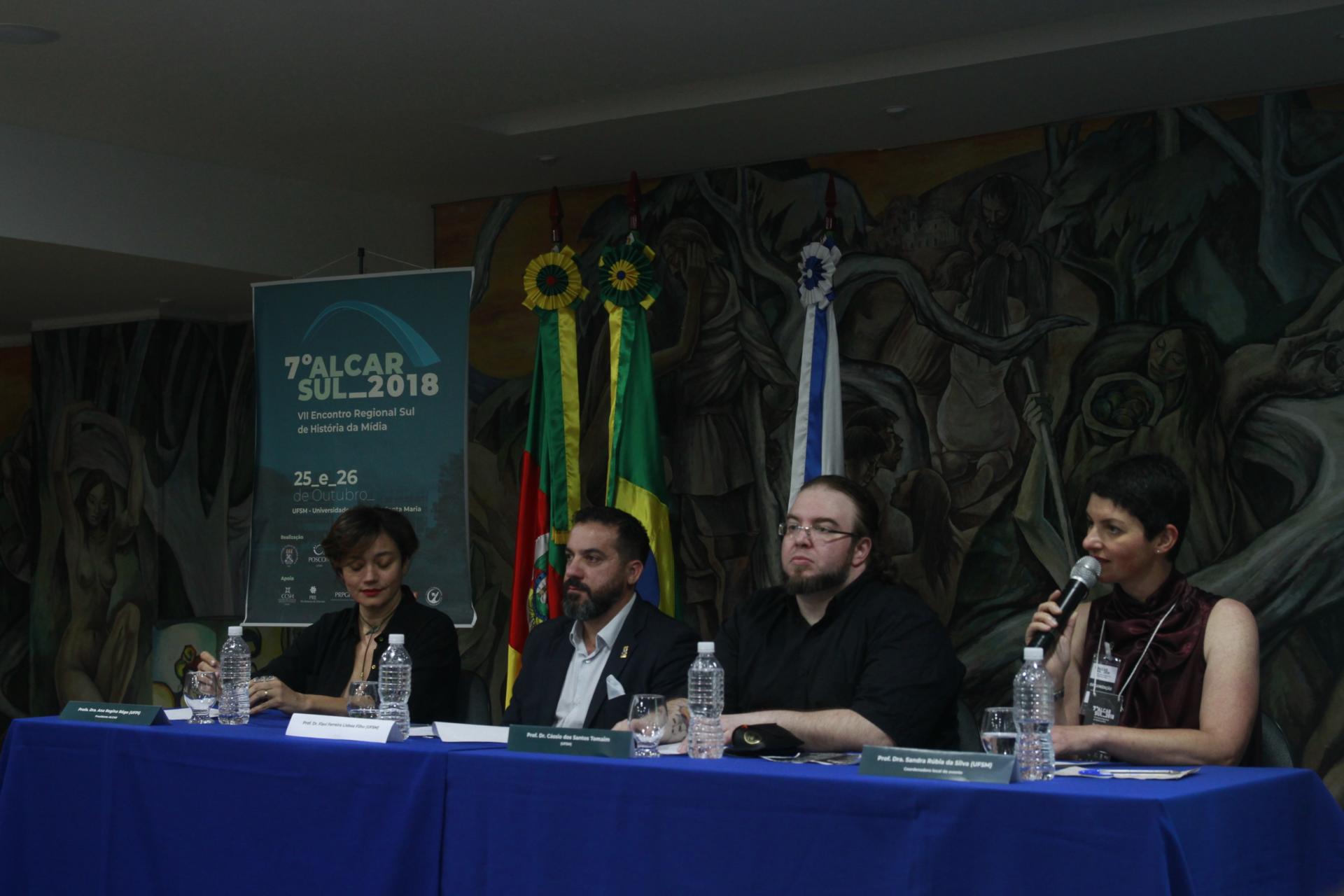 Os quatro componentes da mesa de abertura sentados, perfilados, tendo como fundo o painel do Salão Imembuí, as 3 bandeiras e o banner do evento