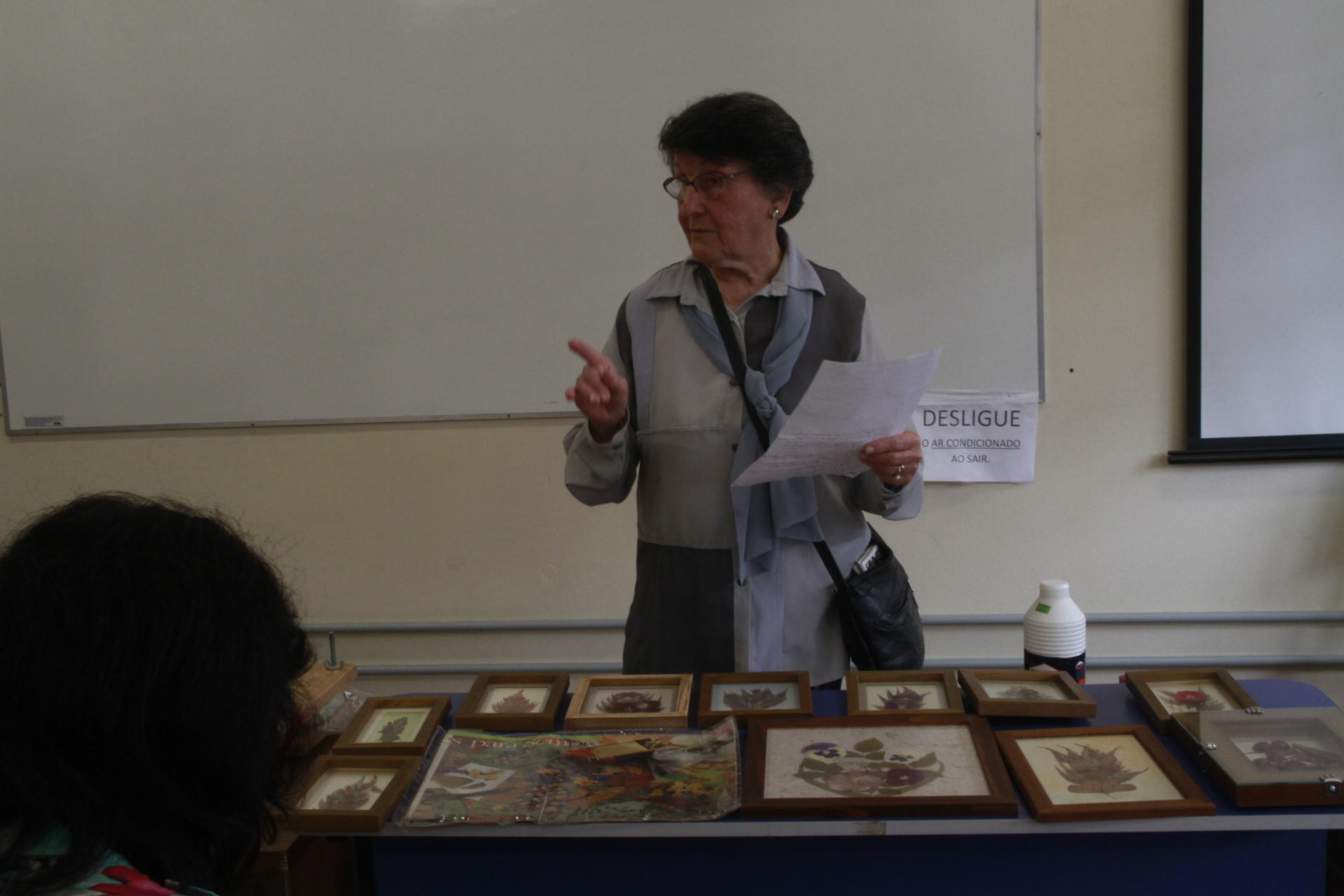 Senhora em pé com folha de papel em uma das mãos e a outra com o dedo em riste. Na mesa várias obras com folhas e flores secas