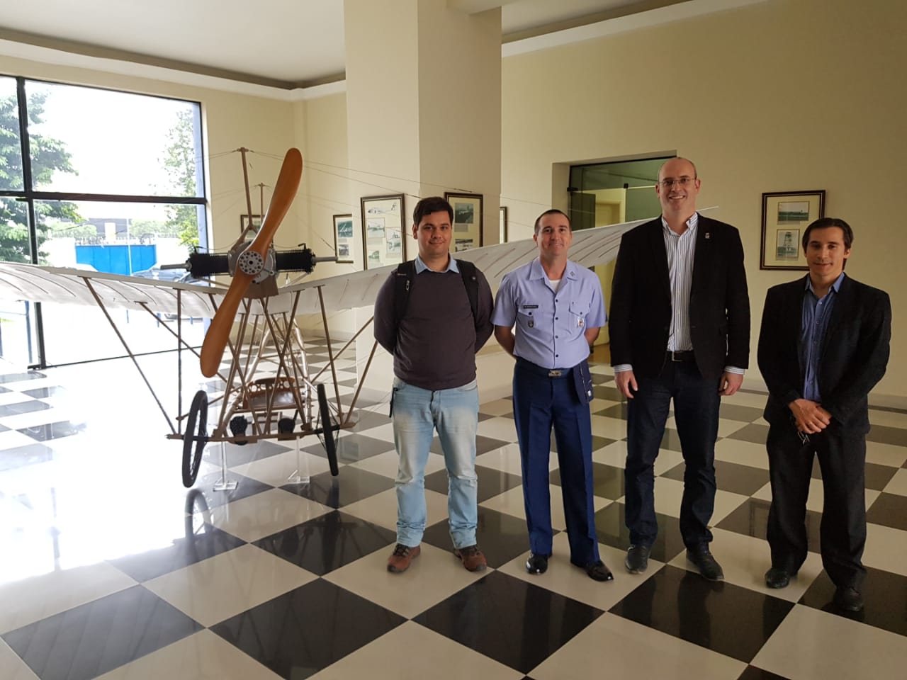 Quatro pessoas estão em um saguão de um prédio em pé, à direita de uma réplica de uma aeronave semelhante ao 14 Bis