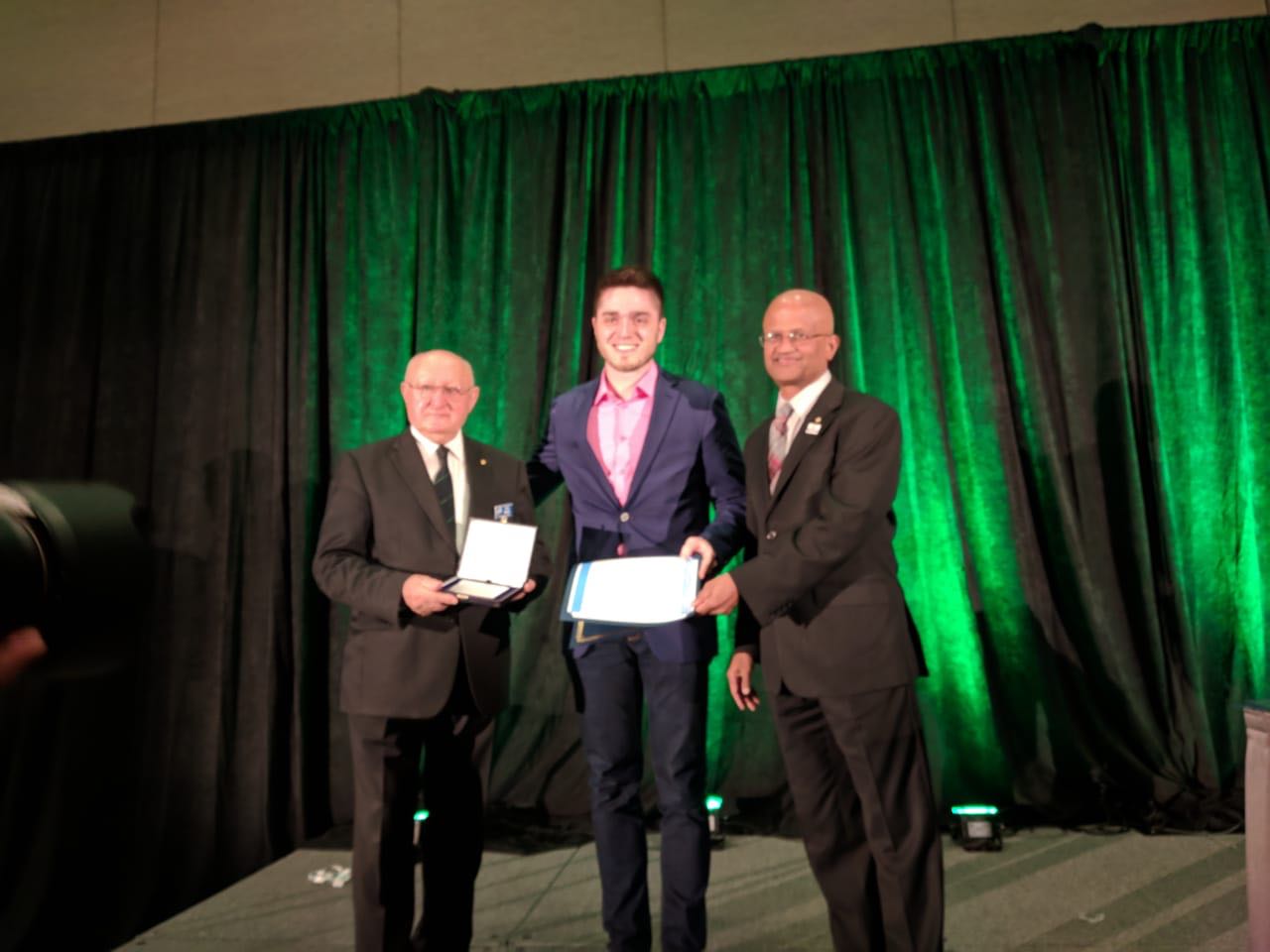 Foto colorida horizontal mostrando o aluno vencedor ao centro com o diploma, e ao lado outras duas pessoas