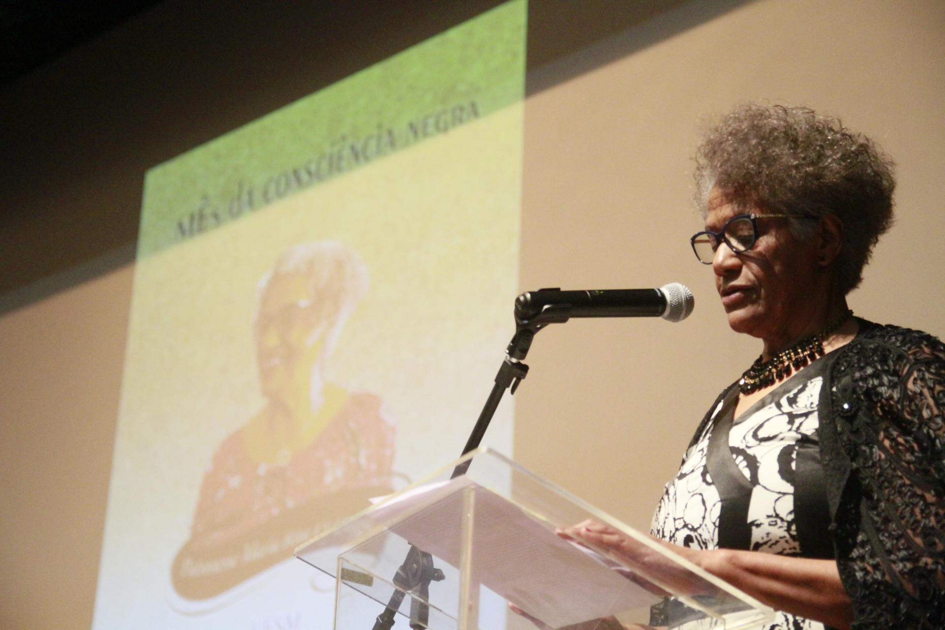 À direita, mulher no púlpito. Ao fundo, imagem projetada com a peça de divulgação do Mês da Consciência Negra