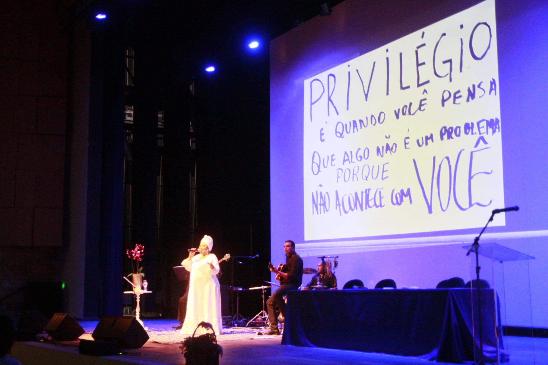 Mulher cantando e atrás, no telão, uma frase sobre privilégio