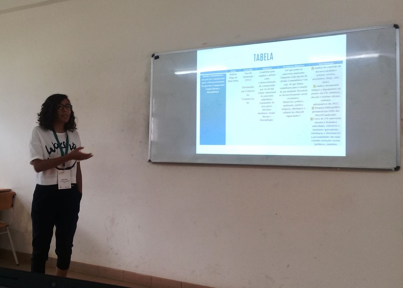 Mulher em pé apresenta trabalho. Ao fundo, projeção de slides com uma tabela