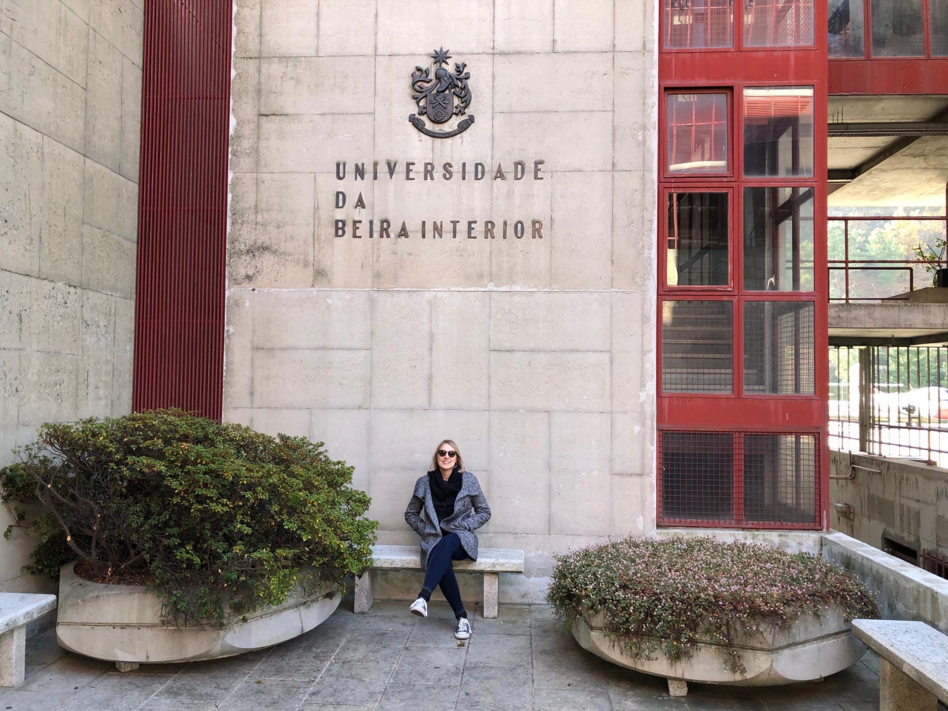 Foto colorida horizontal mostra Carolina sentada em um espaço externo da UBI, à frente de uma parede com o nome e o símbolo da Universidade, e dois grandes vasos de plantas ao lado