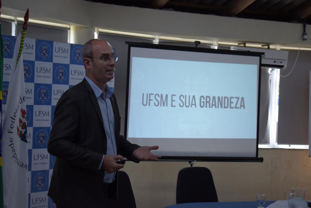 Foto horizontal, em cores, do pró-reitor em pé à frente do público. Ele apresenta um slide sobre a UFSM com a seguinte inscrição: 