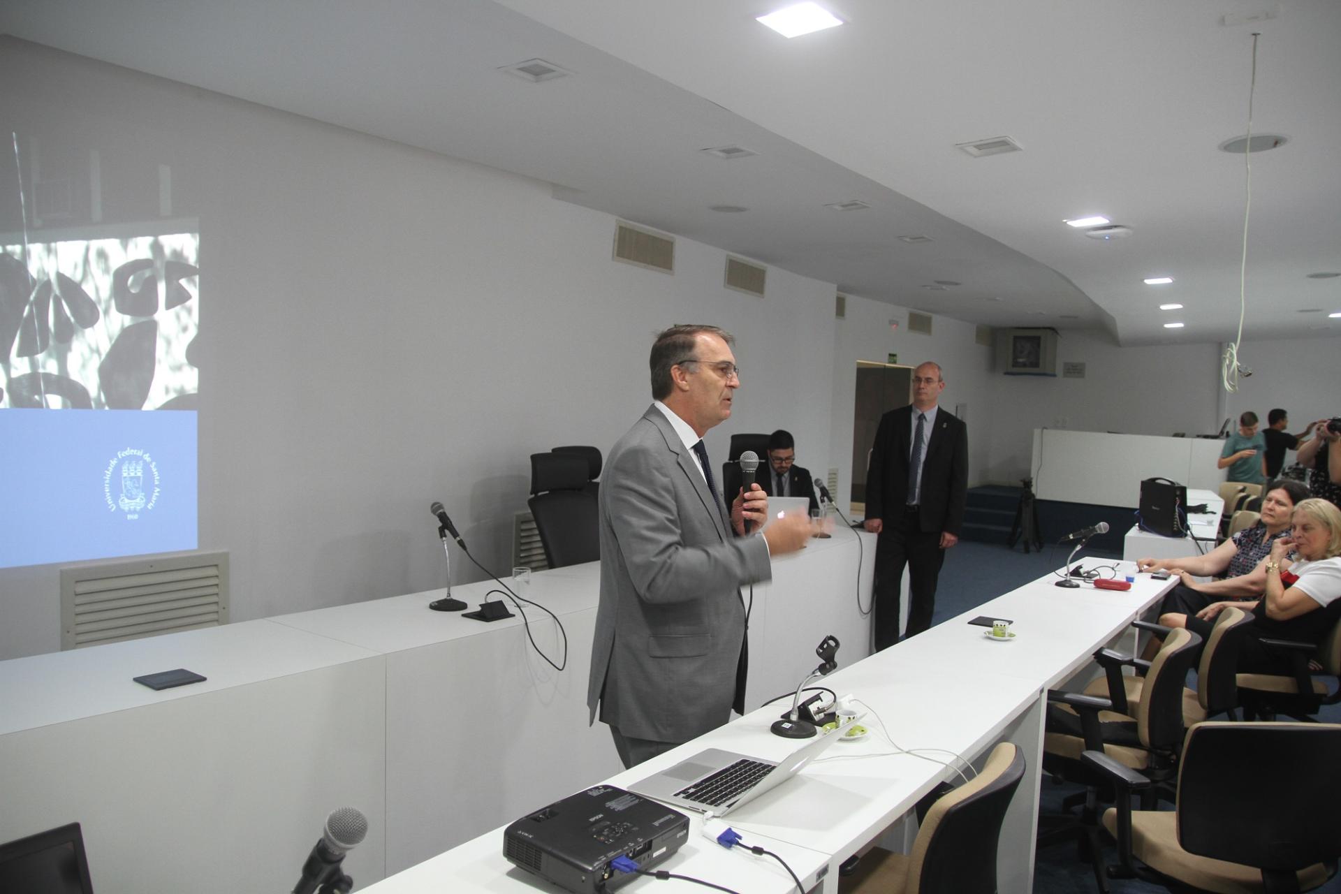 Foto colorida horizontal mostra reitor e vice em primeiro plano, ambos em pé, na Sala dos Conselhos, enquanto o reitor fala ao público