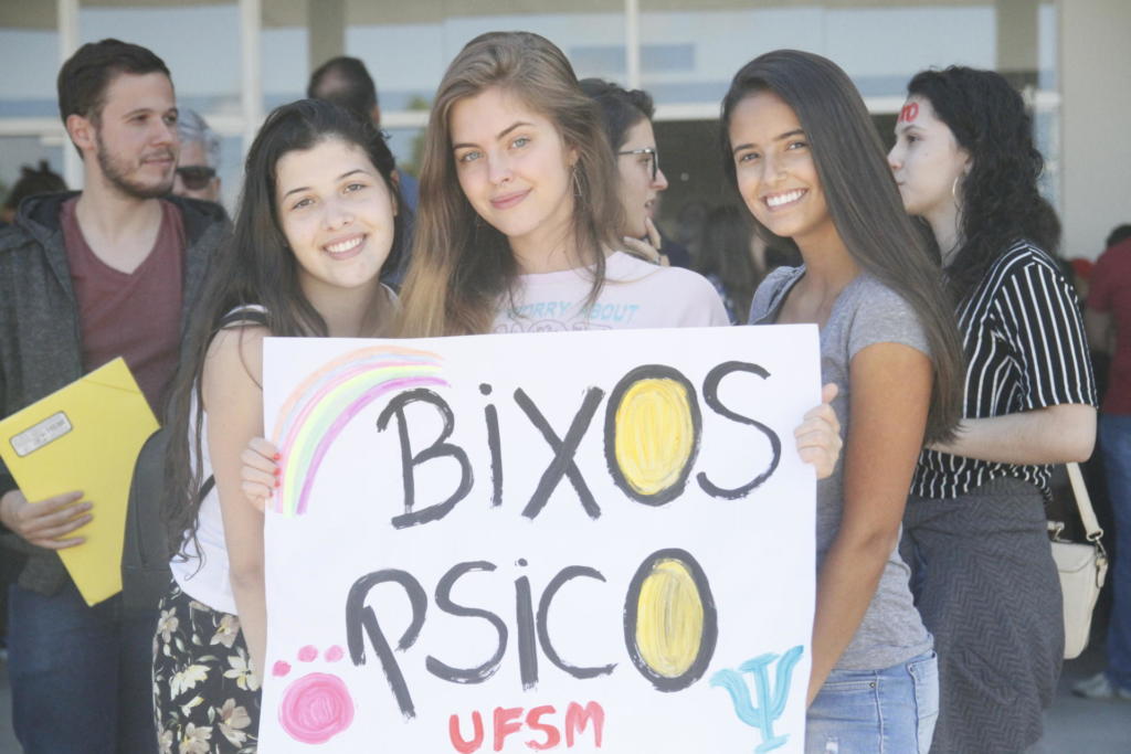 Três estudantes seguram cartaz com o texto "Bixos Psico UFSM"