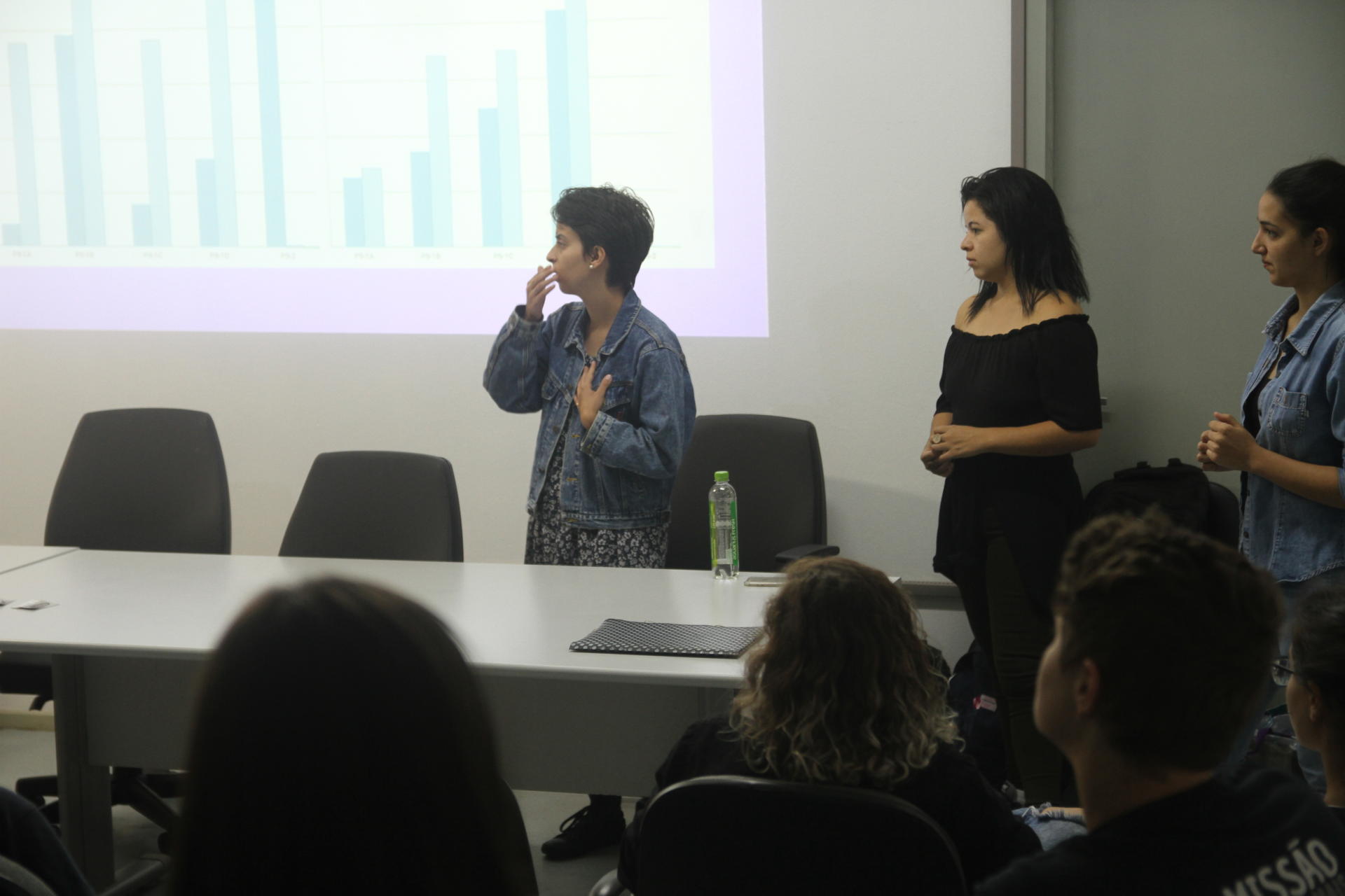 Foto colorida horizontal mostra três alunas, em pé, falando a colegas em uma sala de aula