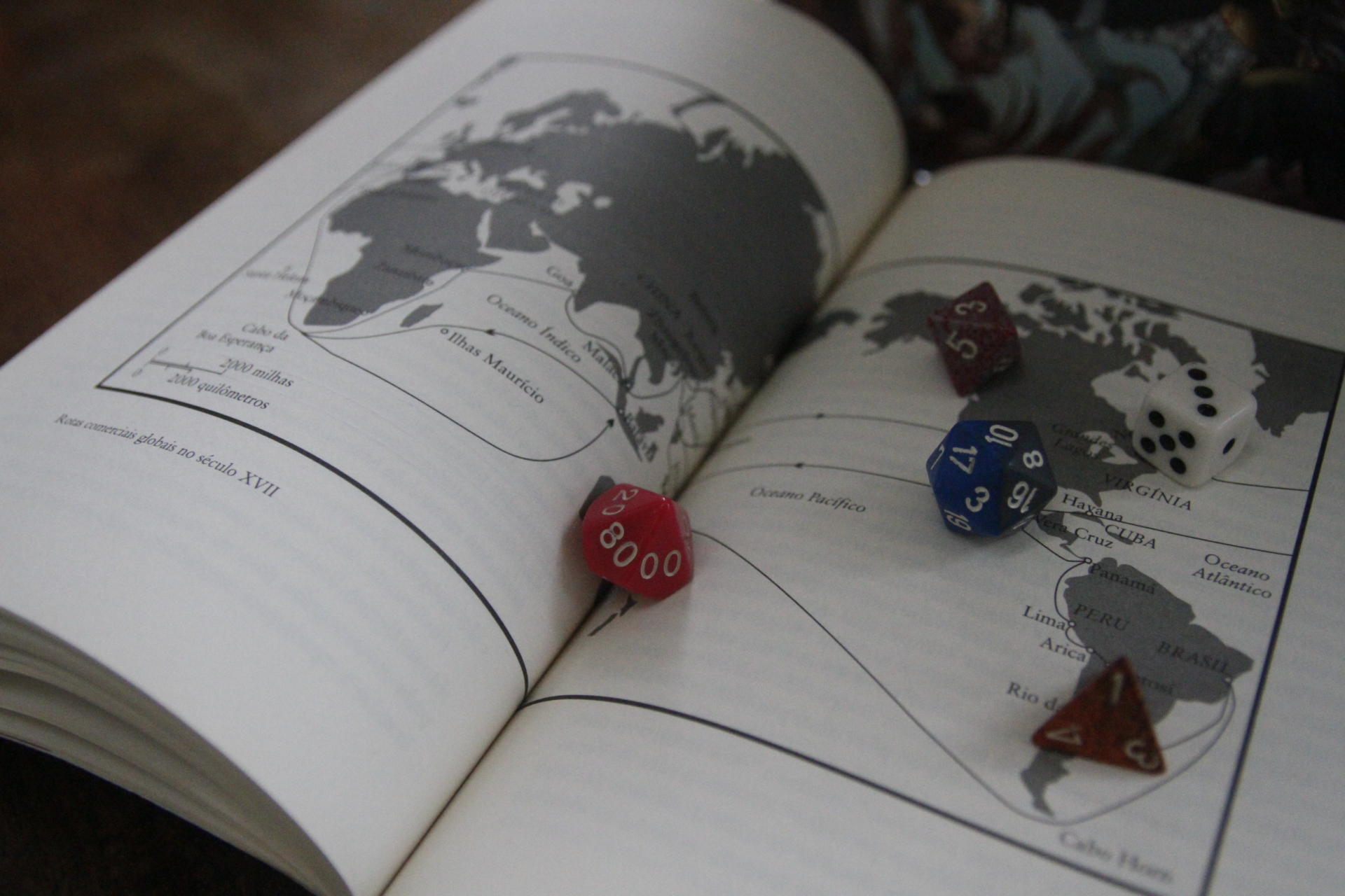 Foto colorida horizontal mostra páginas do livro abertas, com um desenho de mapa em preto e branco ocupando as duas páginas, e sobre o livro cinco diferentes dados