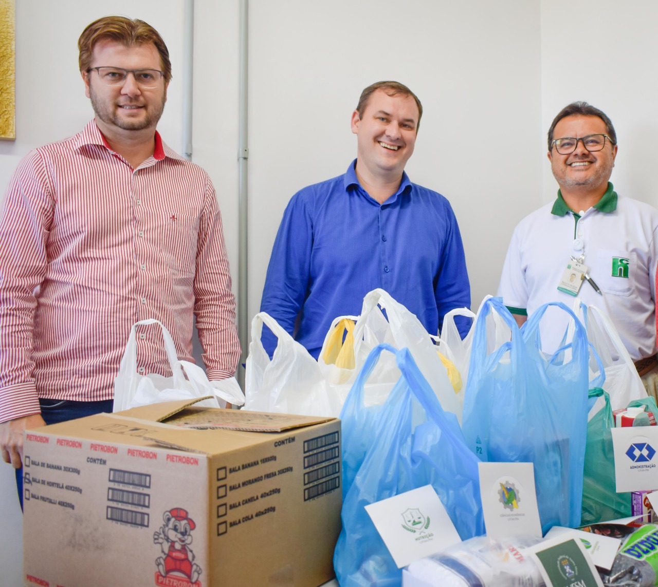 Foto colorida com o diretor do campus ao lado de outros dois homens, representantes do hospital, tendo à frente caixas e sacolas contendo as doações