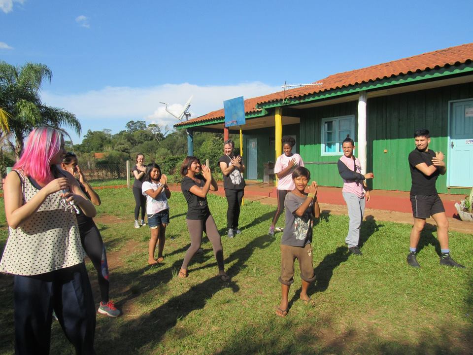 Foto colorida horizontal mostra um grupo de pessoas fazendo movimentos de dança, entre eles crianças indígenas, em um gramado. Ao fundo, casas verdes