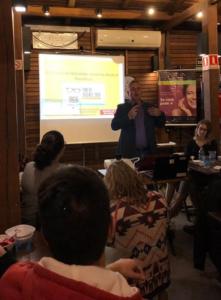 Imagem de professor ao microfone, ao fundo, em um bar com pessoas sentadas