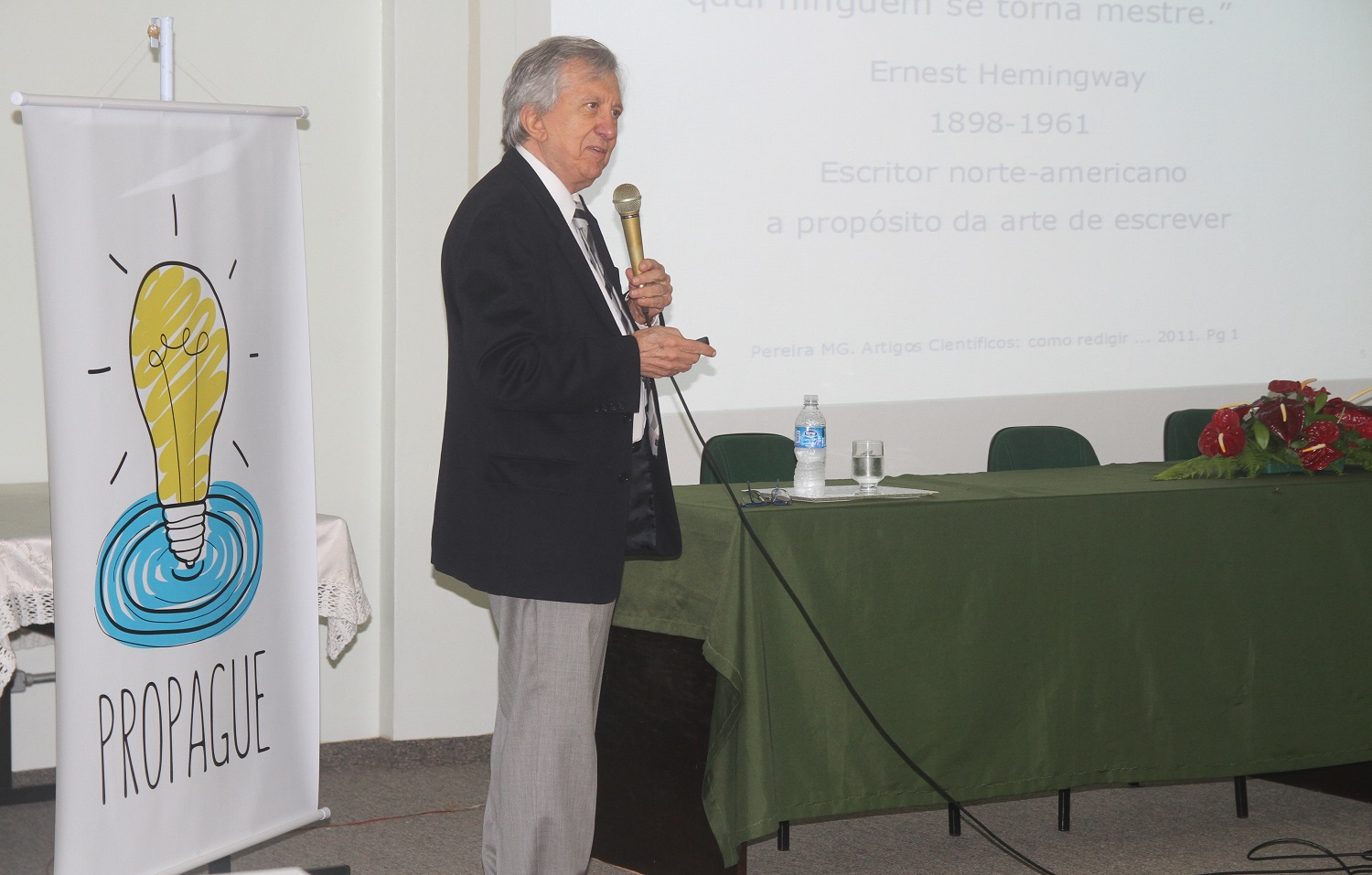 A foto mostra, o palestrante em pé, segurando microfone. À esquerda um cartaz com a marca Propague, que é um lâmpada, e à direita a mesa. 