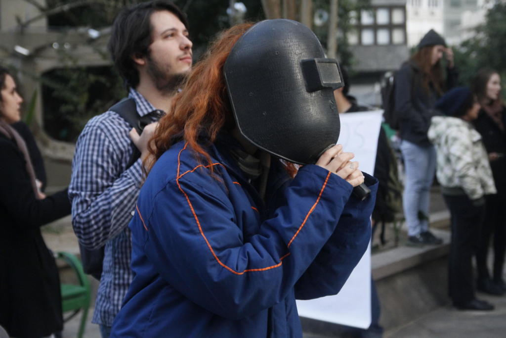 Na foto, em primeiro plano, uma mulher jovem usa uma máscara de soldador. Atrás dela um um homem jovem olha para o céu a olho nu