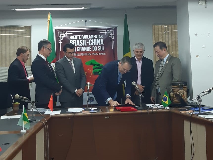 Imagem mostra reitor assinando documento e outros cinco homens, todos em pé. Ao fundo é possível ler cartaz com o seguinte texto "Frente Parlamentar Brasil-China"