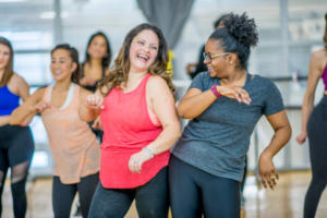 Um grupo multiétnico de mulheres adultas estão dançando em uma academia. Elas estão vestindo roupas de ginástica. Duas mulheres, mais à frente da imagem, estão rindo enquanto dançam juntas.